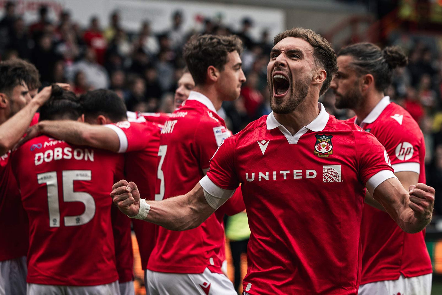 Wrexham AFC players in red jerseys celebrating on a field with spectators in the background