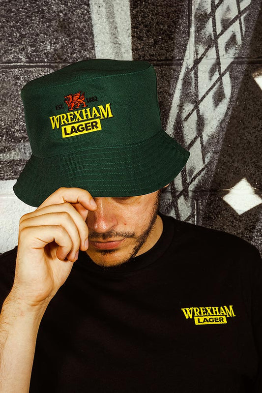Person wearing a green bucket hat and black t-shirt with 'Wrexham Lager' branding against a textured wall.