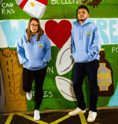 Two people wearing matching light blue Wrexham Lager hoodies in front of a colorful mural.