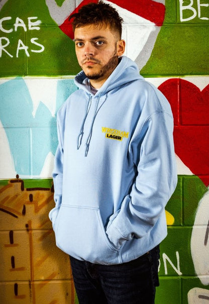 Man wearing a light blue hoodie with a Wrexham Lager logo in front of a colorful mural.