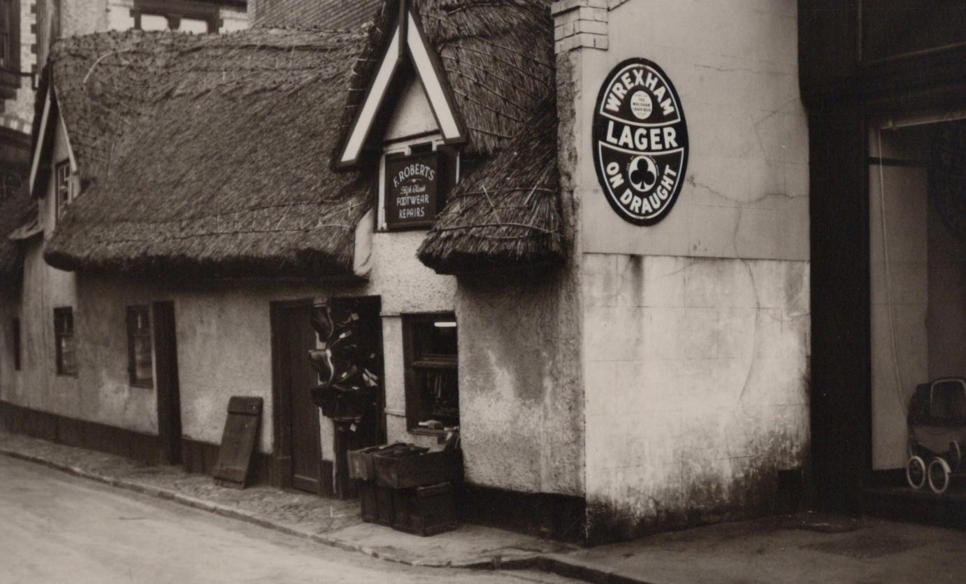 Corner of a building with a thatched roof and a sign for 'Wrexham Lager' in an old-fashioned setting.