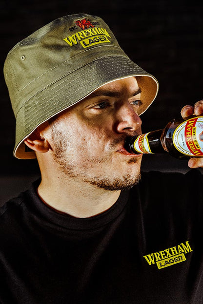 Man wearing a Wrexham Lager bucket hat and shirt, drinking a bottle of Wrexham Lager.