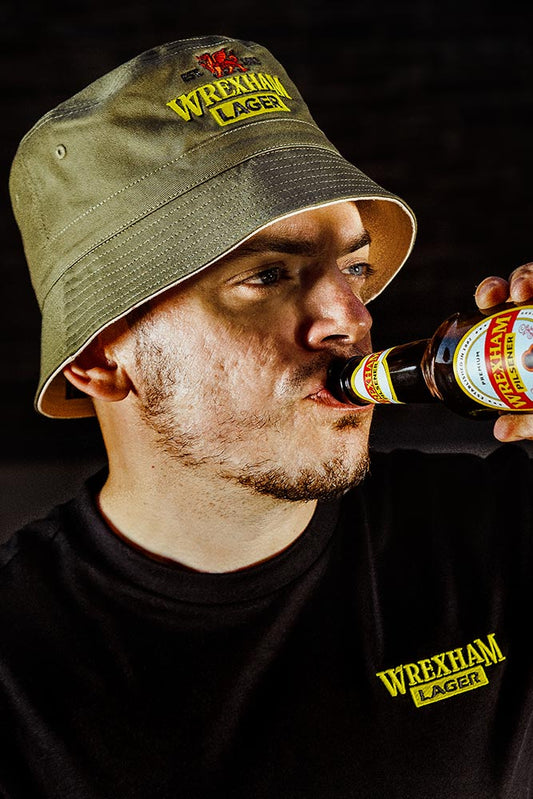 Man wearing a Wrexham Lager bucket hat and shirt, drinking a bottle of Wrexham Lager.