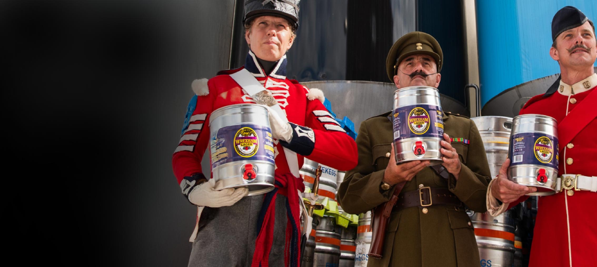 Three Royal Welch Fusiliers in military uniforms holding kegs of Limited Edition Wrexham Lager Royal Welch Fusiliers kegs at the Brewery.