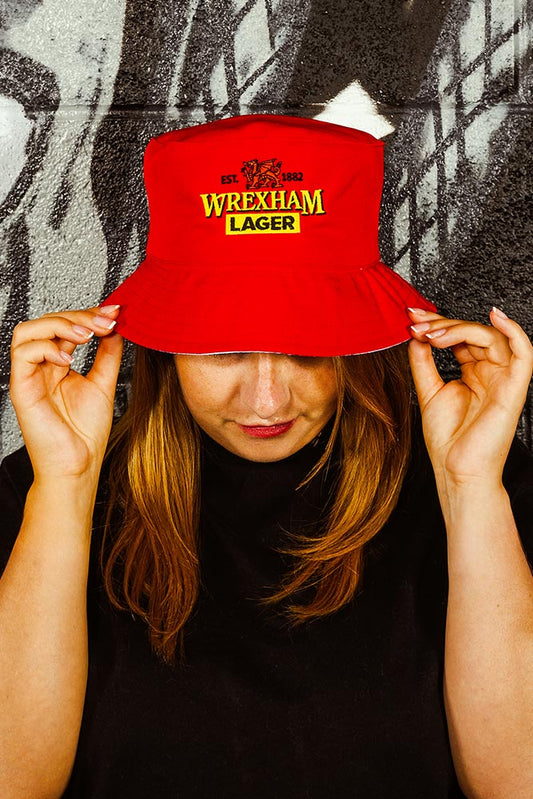 Person wearing a red bucket hat with Wrexham Lager branding against a textured wall.