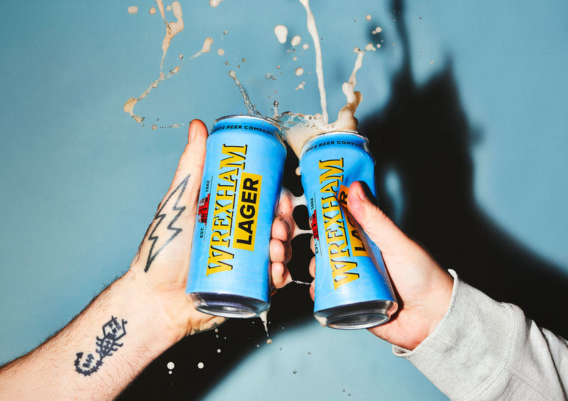 Two cans of Wrexham Lager being held up against a blue background