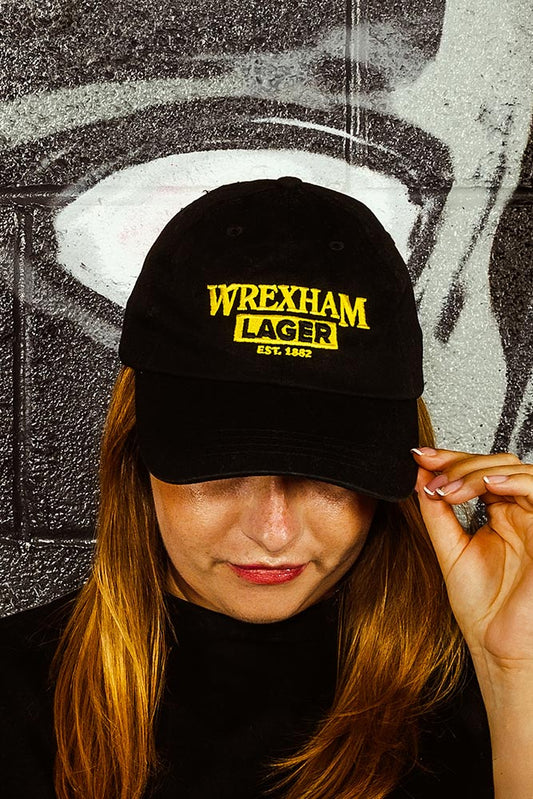 Person wearing a black cap with 'Wrexham Lager' text against a textured wall.