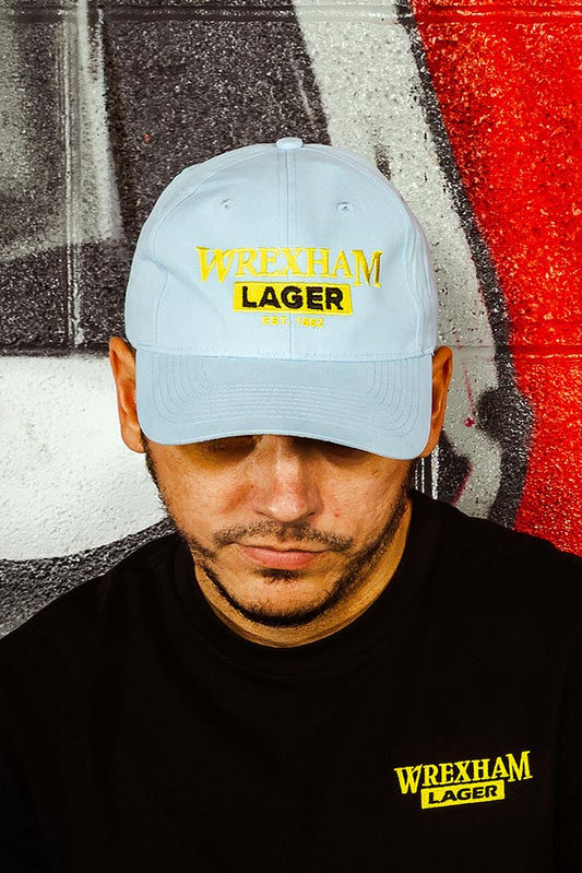 Person wearing a light blue cap and black shirt with 'Wrexham Lager' branding against a graffiti-covered wall.