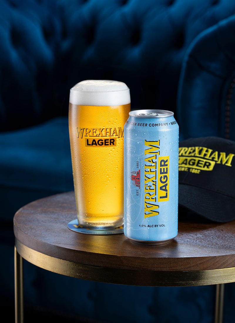 Wrexham Lager Pint and can on a wooden table with a blue background and Wrexham Lager cap