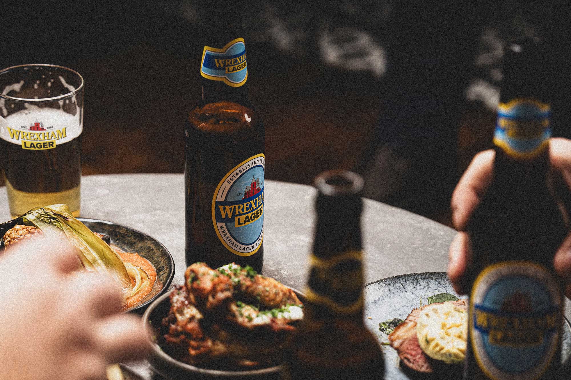 Two bottles of Wrexham Lager and a pint of Wrexham Lager draught on a table with food, one being held by a person.