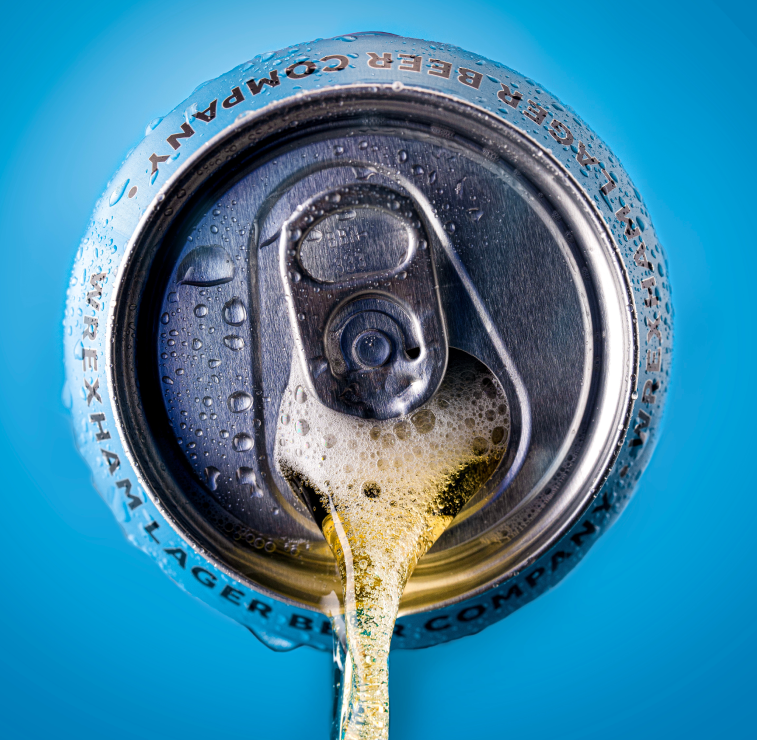 Close-up of a Wrexham Lager can being opened with foam spilling out against a blue background