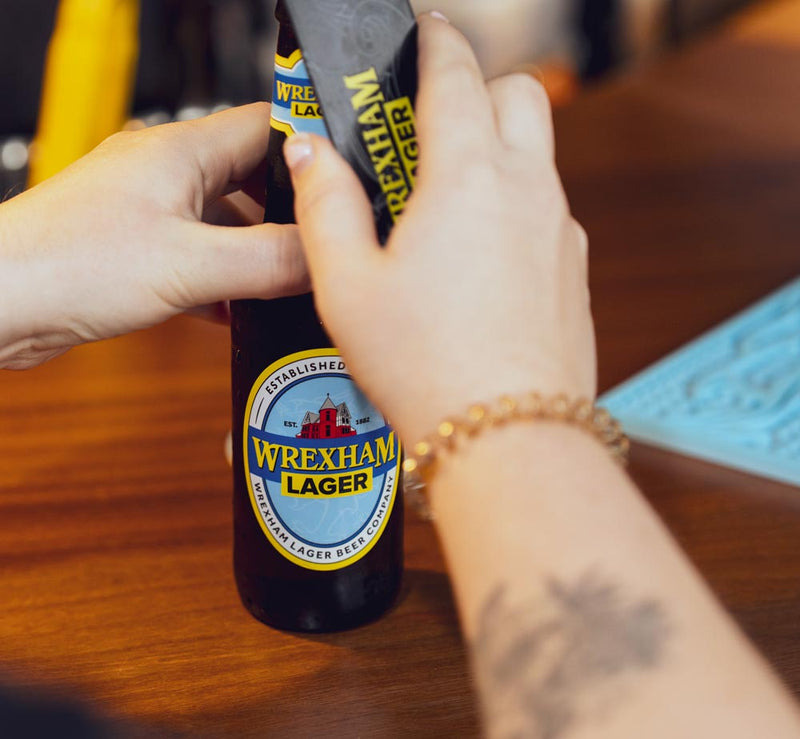 Person opening a bottle of Wrexham Lager with a blurred background