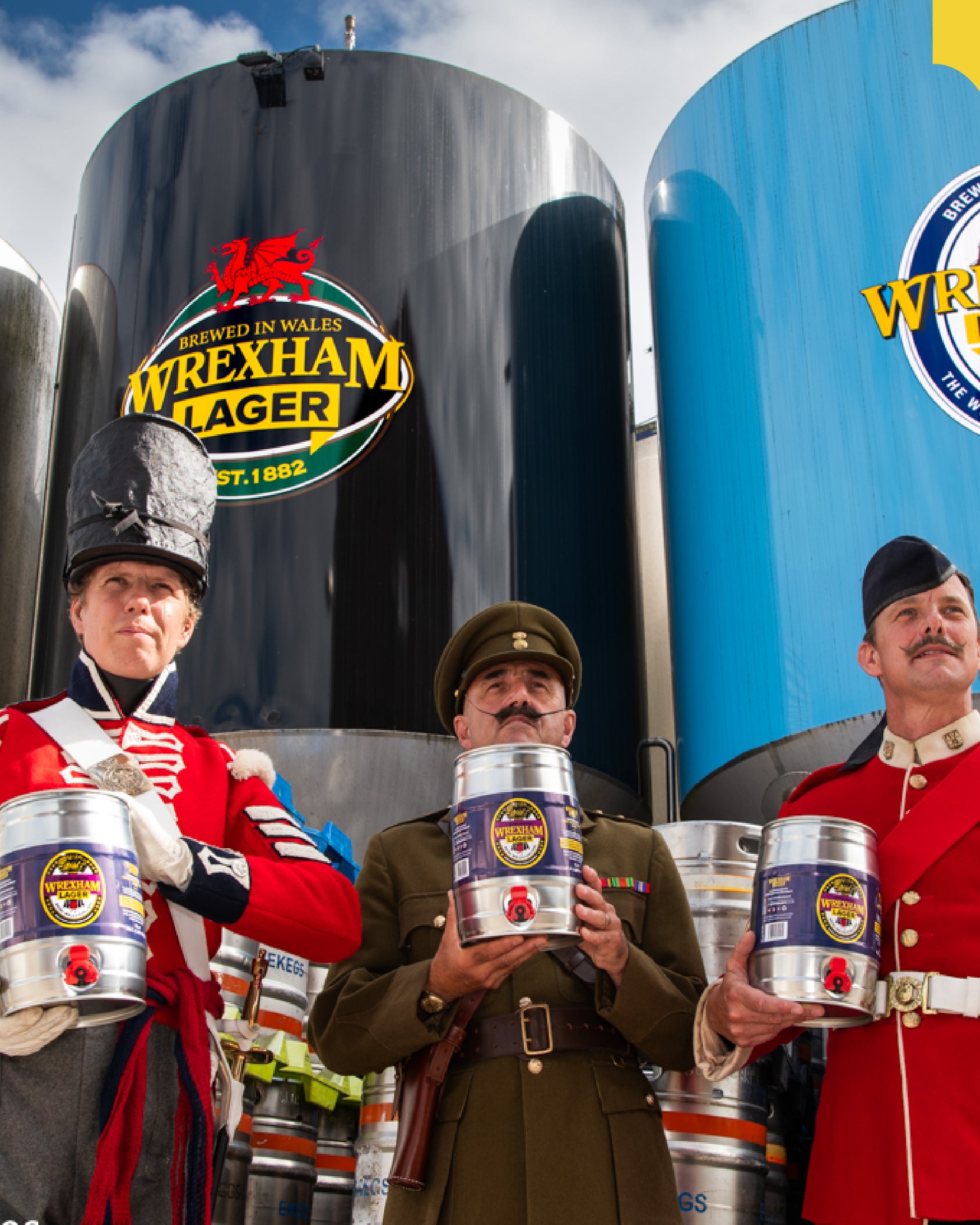 Three men in Royal Welch Fusiliers uniforms holding Wrexham Lager kegs with large storage tanks in the background.