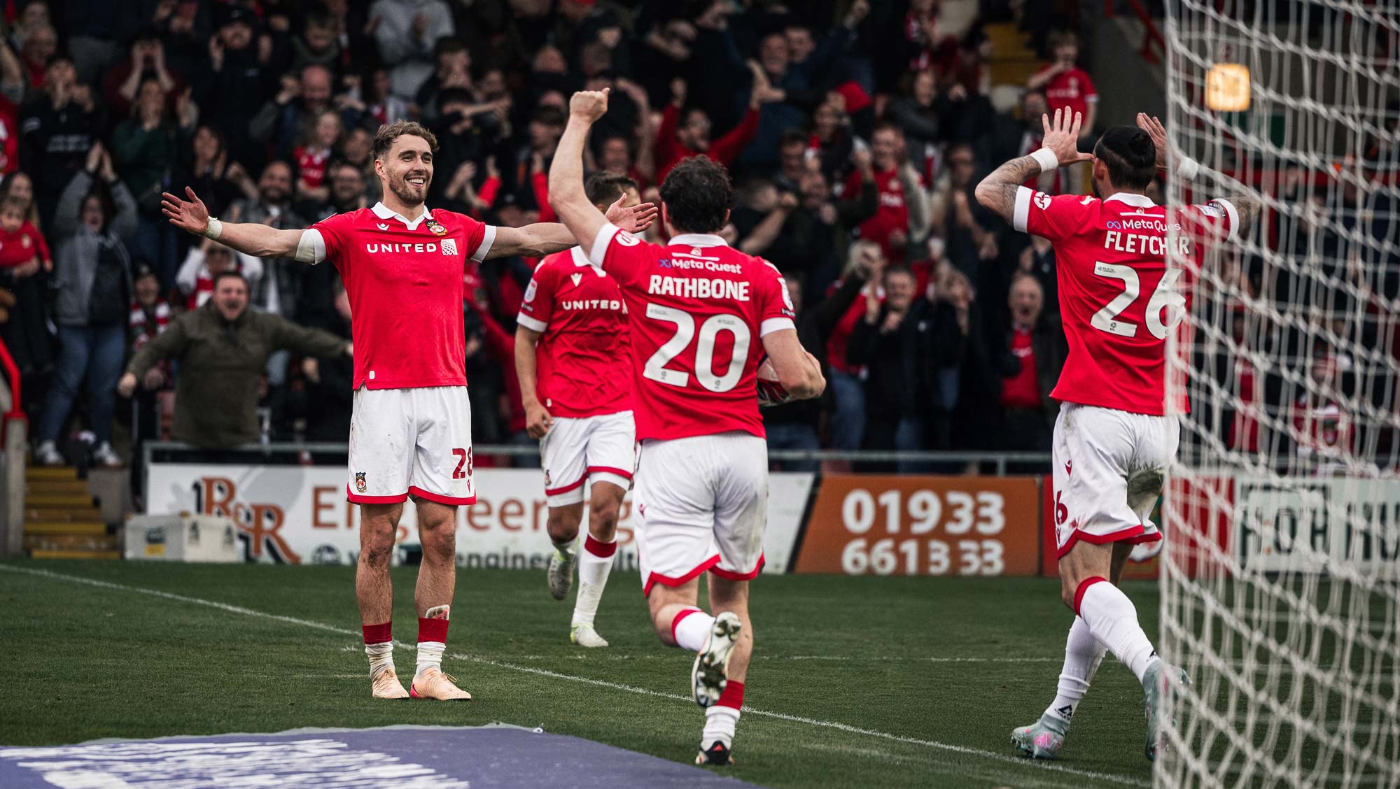 Wrexham AFC players celebrating a goal on a field with spectators in the background.